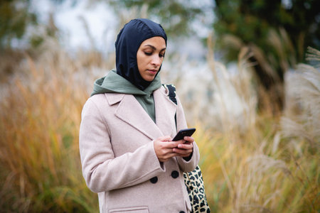 Portrait of young muslim woman in coat, standing outdoor in city park, scrolling smartphone.
