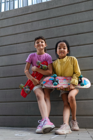Happy friends posing together in city, standing in front of concrete wall, holding skateboards and...