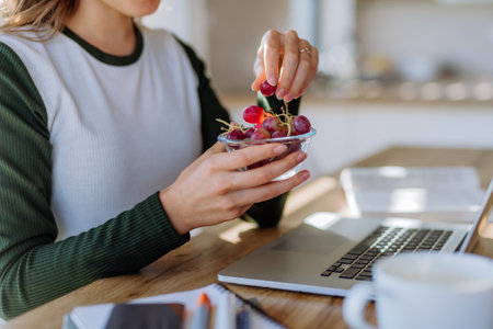 Side view of woman holding bowl with grapes above desk with computer, diary and smartphone. Work-life balance concept.の写真素材