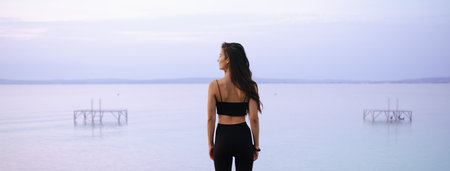 Rear view of young beautiful sportive girl standing by the lake on early morning and contamplating.