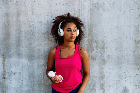 Young multiracial girl in sportswear resting after jogging in city, drinking water from sustainable bottle and listening music in headphones.の写真素材