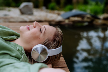 Relaxed woman wearing headphones listening to music sitting on a pier by natureal lake in summerの写真素材