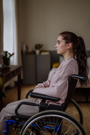 Unhappy teenage girl sitting on a wheelchair and looking out of the window.の写真素材