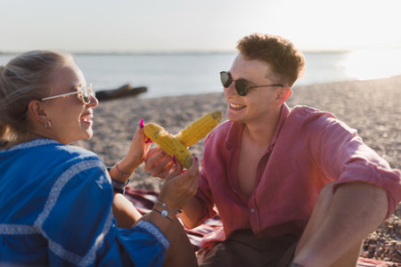 Happy young couple dating together in beach, sitting on a blanket and eating corn. Enjoying holiday time together.の写真素材