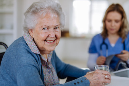 Portrait of senior woman with glass of water, her nurse assistant sitting in background.の写真素材