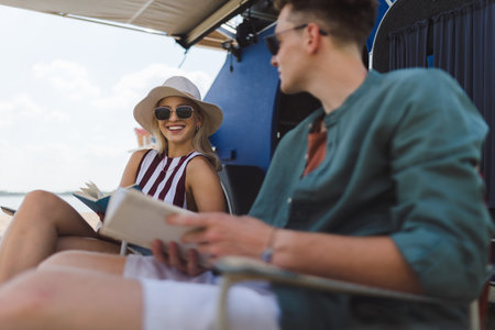 Young couple sitting together in front of van, camping and reading book.