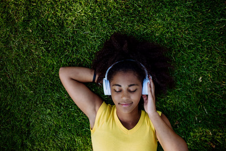 Multiracial girl lying down in grass and enjoying music in headphones, top view.の写真素材