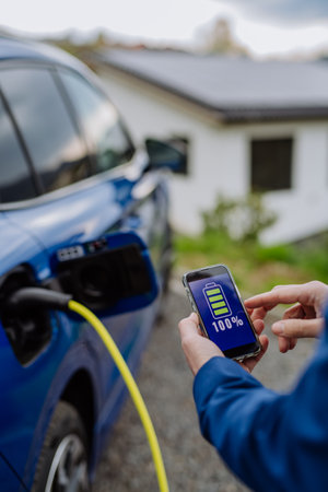 Close up of businessman holding smartphone, checking charging of his electric car.