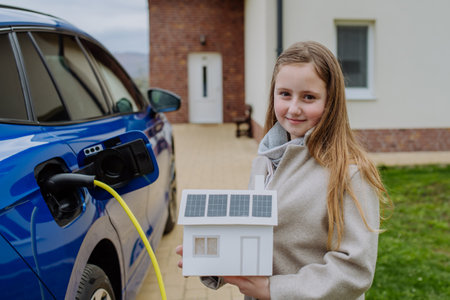 Little girl holding model of paper house with solar panels, waiting for charging electric car.の写真素材