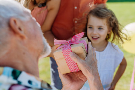 Happy little girl giving birthday present to her senior grandfather at multigenerational family birthday party in summer garden.の写真素材
