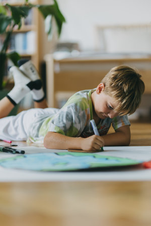 Boy lying on stomach and painting at home with watercolors and tempera paints, creating a model of planet Earth.の写真素材