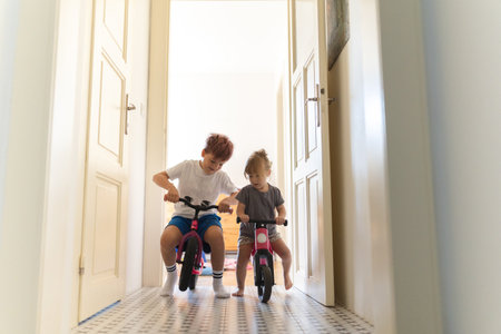 Siblings are riding bicycles indoors, in the hallway of the house.の写真素材