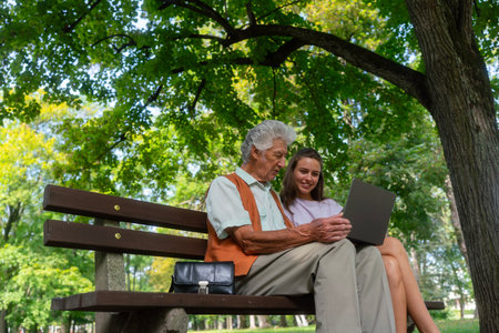 The grandfather and granddaughter are looking at photos on notebook, sitting on a bench in a city park.の写真素材