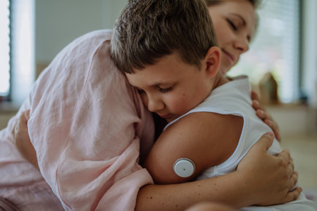 Diabetic boy with a continuous glucose monitor embracing his mother, feeling sad.の写真素材
