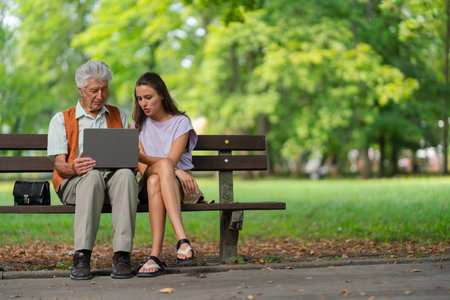 The grandfather and granddaughter are looking at photos on a notebook, sitting on a bench in a city park.の写真素材