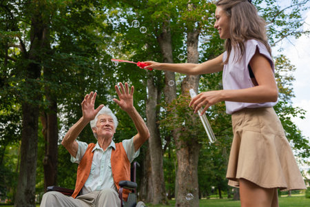 The man in a wheelchair having fun with his caregiver in the park, catching bubbles.