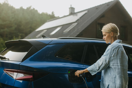 Portrait of a woman charging electric car in front of her house with solar panels on roof. Mother plugging the charger into the charging port. Solar energy and sustainable lifestyle of young family.の写真素材