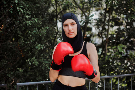 Muslim boxer woman in red boxing gloves and hijab standing outdoors. Woman boxing for relaxation and bolsting mental health. Boxing empowers women. Concept of muslim woman in sport.の写真素材