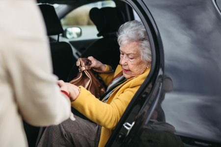 Senior lady getting out of the car, caregiver helping her, holding her hands. Elderly woman has problem with standing up from the car back seat.の写真素材