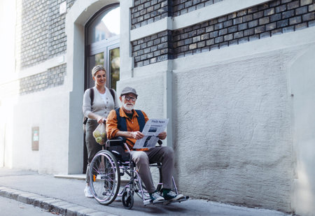 Granddaughter pushing senior man in wheelchair on street. Female caregiver and elderly man enjoying a warm autumn day, going home from shopping.の写真素材