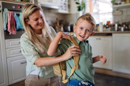 Mother saying goodbye to son before school, helping him to put backpack on back in kitchen.の写真素材