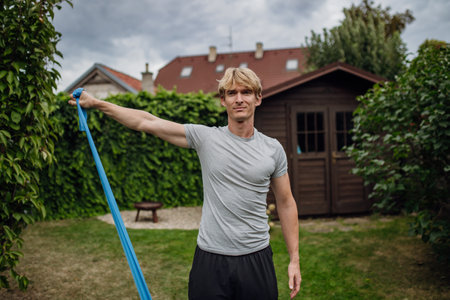 Middle aged man doing strength exercises with resistance band outdoors in garden. Concept of workout...