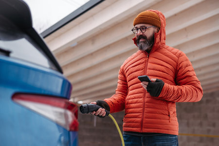 Man charging electric car during cold snowy day, using electric vehicle charging app, checking...