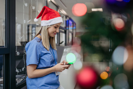 Portrait of beautiful nurse with christmas hat in hospital corridor, holding smartphone. Working in hospital on Christmas day, Christmas Eve. Female doctor working a Christmas shift and cant be with her family.の写真素材