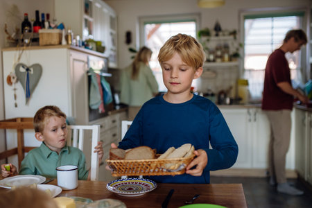 Family preparing breakfast in home kitchen in the morning. Oldest son bringing bread on table.