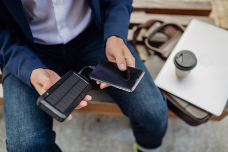 Businessman, freelancer or manager working outdoors in city park. Man with laptop on knees drinking coffee, charging his smartphone with solar phone charger. Concept of working remotely.の写真素材