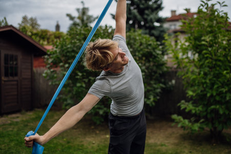 Middle aged man doing strength exercises with resistance band outdoors in garden. Concept of workout routine at home.の写真素材
