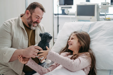 Father sitting beside daughter lying on hospital bed, playing with plush toy. Gift from dad to sick little girl in hospital.の写真素材