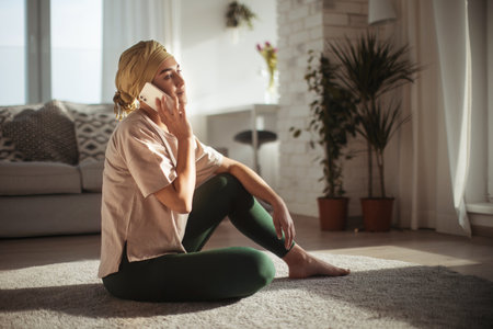 Young woman with cancer sitting on floor, phone calling in her apartment. Strong female patient talking with friend, family on phone, feeling loved.の写真素材