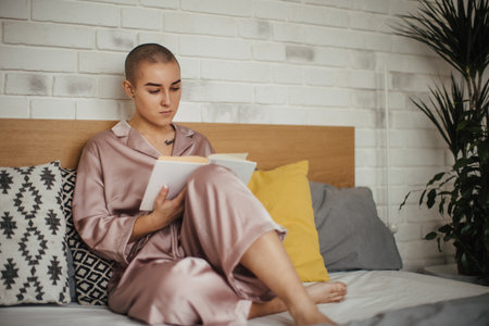 Young woman with cancer reading book, cancer awareness concept. Female cancer patient lying in bed, relaxing at home.の写真素材