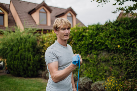 Middle aged man doing strength exercises with resistance band outdoors in garden. Concept of workout...