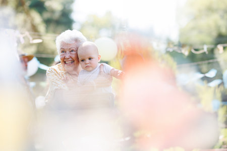 Great-grandmother holding little baby in her arms. Family summer garden party.の写真素材