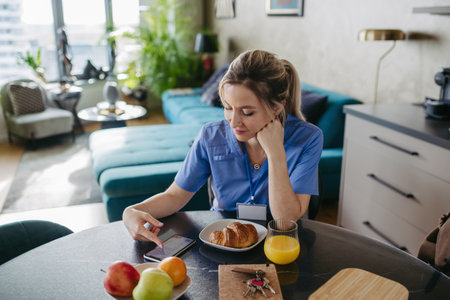 Female nurse or doctor getting ready for work in the morning, scrolling on smartphone while eating breakfast. Work-life balance for healthcare worker.の写真素材