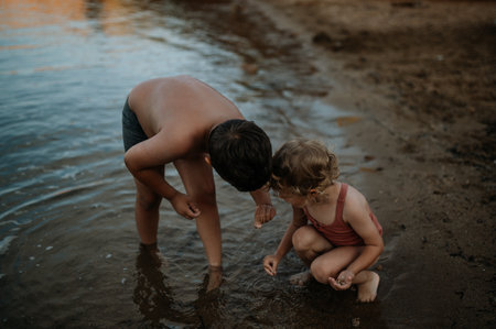 Brother helping sister seraching for shells in sand. Small girl in swimsuit playing at beach, crouching in water.の写真素材