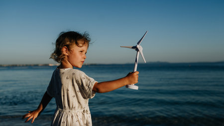 Small girl with blonde hair standing at beach, holding wind turbine model in hand, sea wind spinning...
