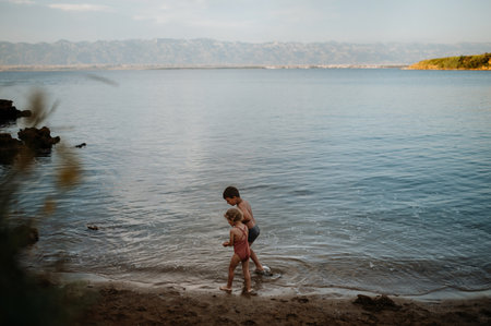 Brother helping sister seraching for shells in sand. Small girl in swimsuit playing at beach, crouching in water.の写真素材