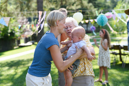 Great-grandmother holding crying little baby in her arms. Family summer garden party.