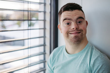 Young man with Down syndrome sitting by window, looking at camera.