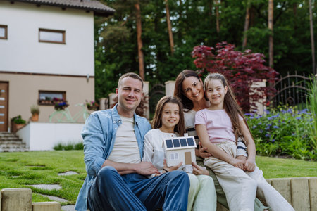 Happy family holding model of house with solar panels on roof and wind turbine. Concept of renewable, green energy and sustainable lifestyle.の写真素材