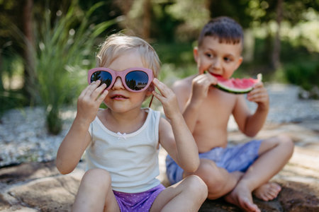 Little chidren sitting near lake and eating watermelon on hot sunny day during summer vacation.の写真素材