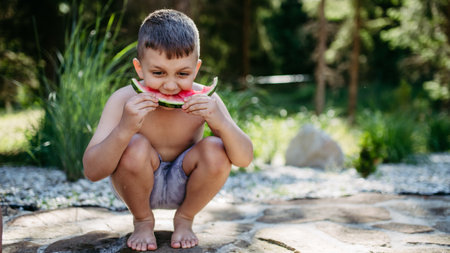Little boy sitting near lake and eating watermelon on hot sunny day during summer vacation.の写真素材