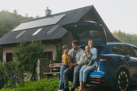 Family with electric car standing in front their house with solar panels on roof. Solar energy and sustainable lifestyle of young family. Concept of green energy and sustainable future for next generations.の写真素材