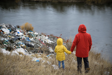 Little girl with father standing on river shore looking at piles of waste, debris on riverbank. Water and environmental pollution, eco activism concept.の写真素材