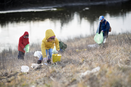 Little girl among eco activists picking up litter, along riverbank in nature, environmental...