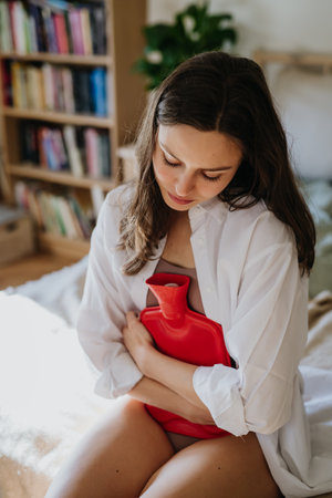 Woman at home suffering from menstrual pain, having cramps. Woman warming lower abdomen with a hot water bottle, endometriosis, and conditions causing pain in tummy.の写真素材