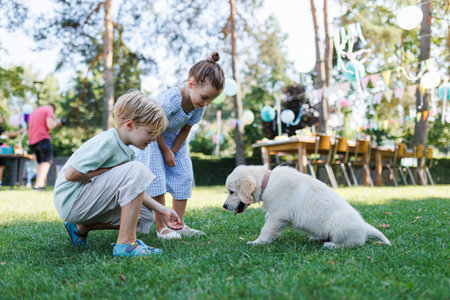 Children playing with a small puppy at a family garden party. Portrait of little boy and girl on grass with Golden retriever puppy.の写真素材